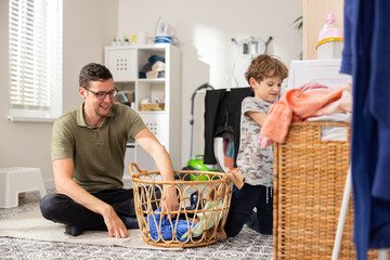 The boy helps his dad put dirty clothes in the washing machine, they take things out of the wicker basket, the man smiles with pride and gratitude that the child is helping him.
