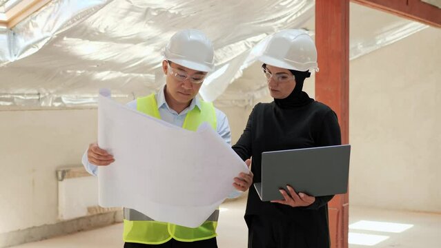 Muslim female investor wearing a hijab protective helmet and goggles talking to Asian engineer. Arab female holding laptop showing investment project discuss investment in the construction business.