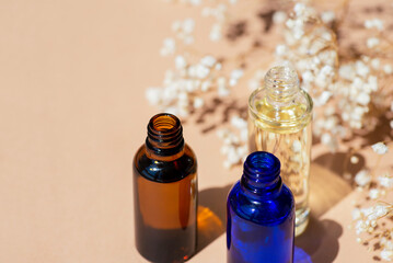 Open amber, blue and transparent bottles without cap with serum or essential oil with white flowers. Beige background with daylight and beautiful shadows. Beauty concept for face and body care