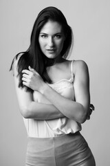 Naklejka premium Fashion and make-up concept. Beautiful woman fashion studio black and white portrait with long dark hair with seductive look and pose with crossed arms. Model wearing white blouse and velvet trousers