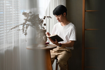 The portrait of young smart Asian male reading a book next to the sunshade window over blurred...