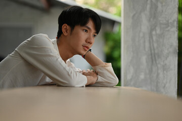 The portrait of lonely young Asian male sitting hence to rest the chin on the hand and looking thoughtful over the concrete building background. Asian man portrait. Thai with native dressing concept.