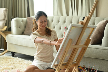 A young Asian woman is holding a paint palette and paintbrushes while happily painting on canvas at...