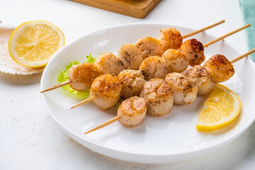 Grilled scallop skewers in spices until golden brown appetizing crust in a white plate close-up with a piece of lemon.