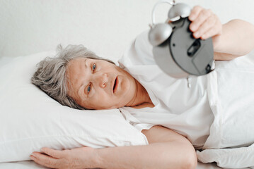 Senior woman with surprised expression holding an alarm clock while lying in bed. Overslept elderly woman looking shocked at alarm in bedroom in morning