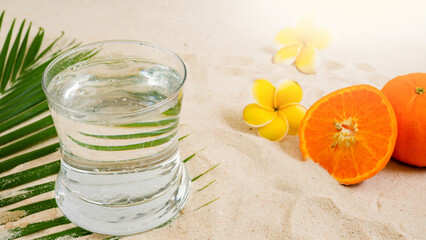 Close Up Glass Sparkling Water On The Sand Beach Against Orange And Plumeria Flowers.