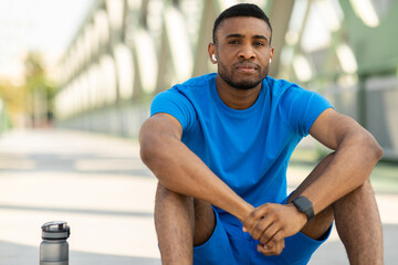 Fototapeta na wymiar Relaxed african american sportsman with wireless headphones sitting on the floor and listening to music