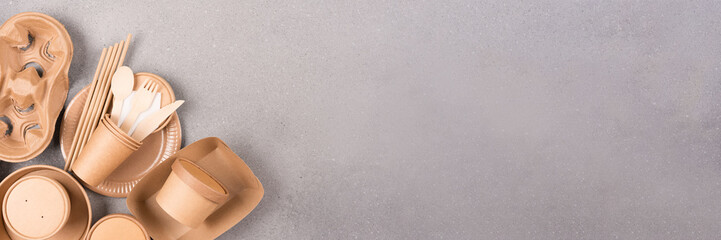 Banner with eco paper tableware and utensils - paper cups, containers and wooden bamboo cutlery over gray concrete background with copy space. Sustainable food packaging concept