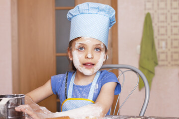 5 year old girl in blue dress and dressed as chef in kitchen with face smeared with flour