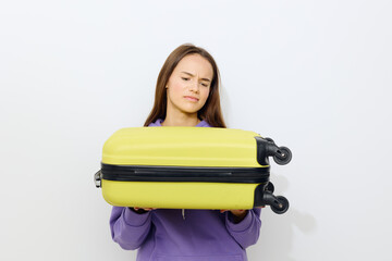 a disgruntled beautiful woman stands on a white background in a purple tracksuit holding a suitcase from below with both hands frowning a little