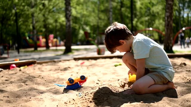 a five-year-old boy walks on a playground in a summer park. A child digs sand with a shovel