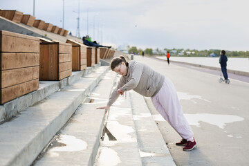 overweight European teenage girl in tracksuit does push-ups on stairs concrete embankment, on outskirts of city. Sports exercises and teenagers, overweight teenagers,