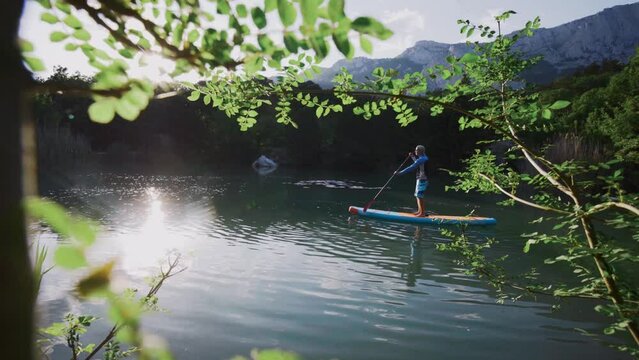View through bush on shore of mountain lake to SUP surfer rows with orange paddle standing on blue board sailing along calm water with forest and bright sunlight reflections