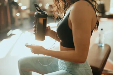 Poster Cropped picture of a strong sportswoman sits in a gym, drinks protein drink and using a phone while drinking protein drink and taking a break. © chika_milan