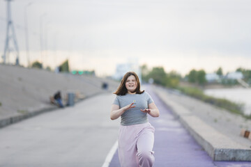 Overweight European teenage girl in tracksuit warms up, runs on violet floor concrete embankment,...