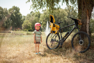 Beautiful little girl wearing pink protective helmet stands next to a bicycle with a back child bike seat. Safety while cycling with a kid in the nature