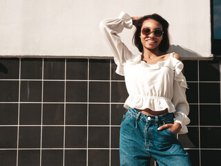 Portrait of young beautiful black woman. Smiling hipster model in white jeans clothes. Sexy carefree female posing on the street background. Cheerful and happy outdoors. In sunglasses. At sunset