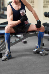 Fototapeta na wymiar Cropped shot of an athletic man in a sportswear and blue knee socks with a color print. He is sitting down on a bench with a dumbbell in his hand. The man in golfs is isolated on the gym background.