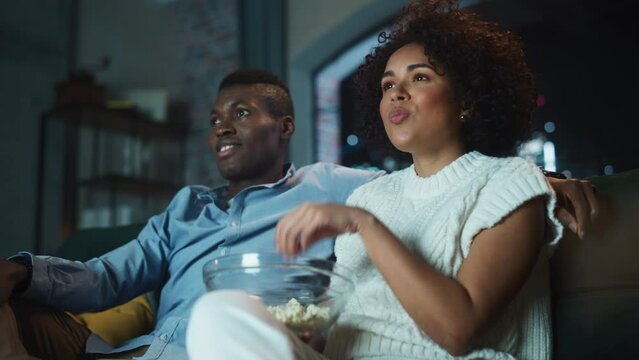 Black Couple Watching Comedy Movie on TV, Eat Popcorn while Sitting on Couch in the Apartment Late at Night. Laughing Boyfriend and Girlfriend Enjoying Funny TV Series Together at Home. Low Angle Shot