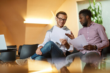 Cheerful optimistic young multi-ethnic men sitting on chairs in conference room and analyzing papers together