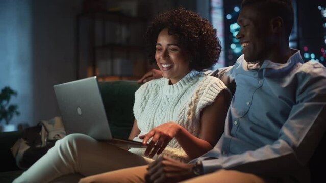 Multiracial Girlfriend and Boyfriend Sitting on a Sofa with Laptop on the Knees, Working Remotely. Woman Sharing her Opinion with Black Man During the Evening