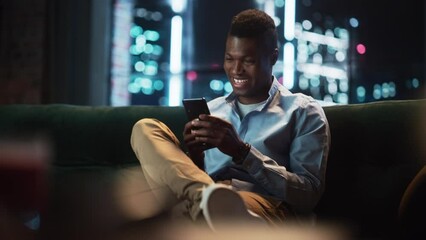 Excited Black African American Man Using Smartphone while Sitting on a Sofa in Living Room. Happy Man Smiling at Home and Chatting With Friends Over the Internet. Using Social Networks Concept