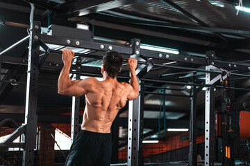 Fototapeta na wymiar Back view of muscular sportsman doing pull ups on horizontal bar at gym. Sports trainer in gym workout exercising. Fit and athletic man having naked torso, wearing in black shorts..