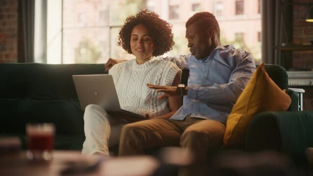 Cute Black Couple in Love Use Laptop Computer, Shopping on Internet, Using Social Media. Boyfriend and Girlfriend Have Fun, Laughs, while Sitting on the Couch in the Cozy Apartment. Female Empowerment