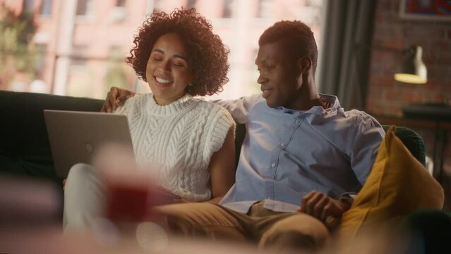 Free Time Together. Half-Length Portrait of Black Couple Relaxing on Couch at Home while Looking in Computer. Happy Boyfriend and Girlfriend Talk and Laugh Using Laptop