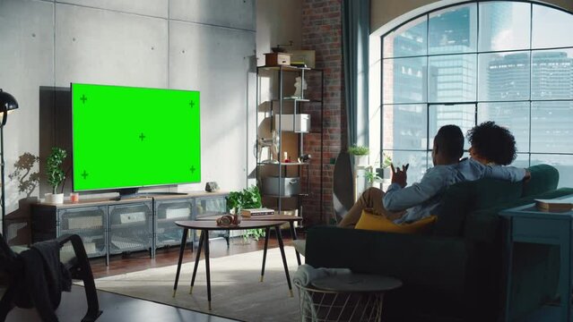 Black Man with Remote Controller at Hand Pointing at Chroma Key Display and Talking to his Wife During the Watching TV at Sofa. Young Couple Spending Time at Home