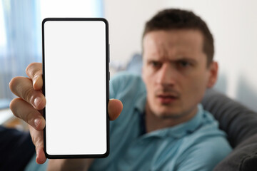 Upset man showing smartphone with blank white screen. Mobile phone in focus with empty screen for mock up, copy space. Out of focus is angry man sitting on sofa at home.
