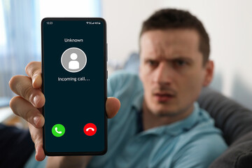 Upset man holding a smartphone with unknown caller displayed on screen. Unknown caller telephony concept. Incoming call at mobile phone. Prank caller, scammer or stranger, fraud, cybercrime. - obrazy, fototapety, plakaty