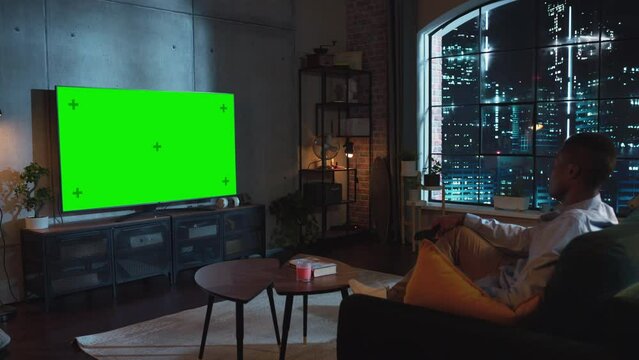 In Loft Living Room African American Man Sitting on a Couch, Turns On TV with Green Screen Mock Up Display with a Controller. Male Enjoys Leisure Time at Home. Night Shot