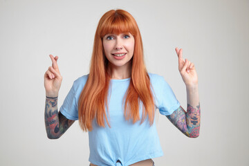 Indoor portrait of young ginger female posing over white wall crossed her fingers with excited...