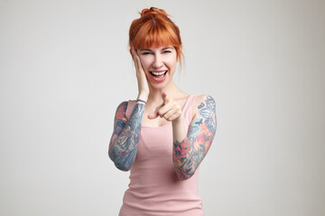 Indoor portrait of young ginger female posing over white wall points with a finger into camera with...