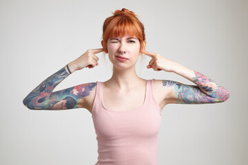 Indoor portrait of young ginger female posing over white wall plug her ears with a fingers