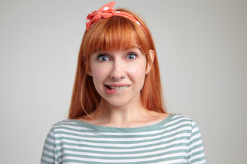 Indoor portrait of young ginger female posing over white wall looking into camera, biting her lip with excited facial expression - obrazy, fototapety, plakaty