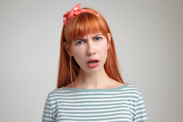 Indoor portrait of young ginger female posing over white wall looking into camera with shocked and confused facial expression