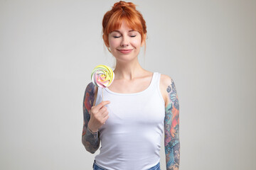 Indoor portrait of young ginger female posing over white wall holding big colorful candy and smiles