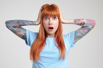Indoor portrait of young ginger female posing over white wall looking into camera with shocked facial expression