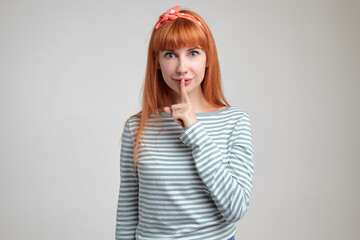 Indoor portrait of young ginger female posing over white wall showing silence gesture into camera