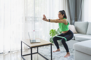 Fototapeta na wymiar Young woman wearing green sport wear doing squat and body weight exercise with resistance band in the morning at home and using online lesson on laptop with personal trainer before starting work.