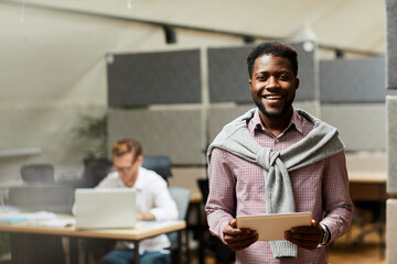 Happy handsome young African-American intern of IT company holding digital tablet and looking at camera in office