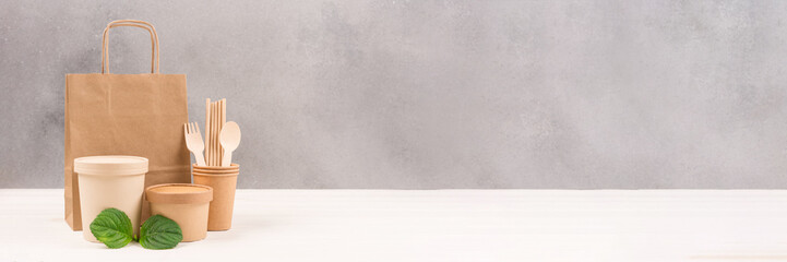 Paper food containers, bag, cups, drinking straws and wooden cutlery set against gray wall background with copy space. Banner for sustainable food packaging concept. Eco tableware