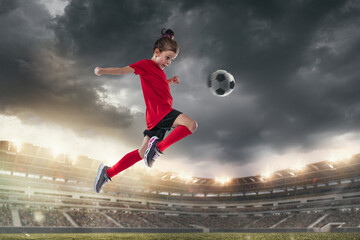 Portrait of little girl, football player in red uniform hitting ball with knee in a jump isolated over open air stadium.
