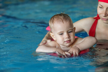 Fototapeta na wymiar Small baby boy learns to swim on swimming board in pool with support of mother.