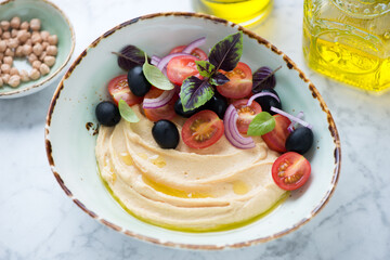 Bowl of hummus served with cherry tomatoes, black olives, red onion and basil leaves, middle closeup on a light-grey marble surface
