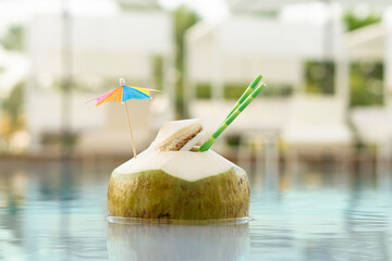 Fresh  green coconut drink with paper straw  and rainbow umbrella  swimming in  pool  water  on ...
