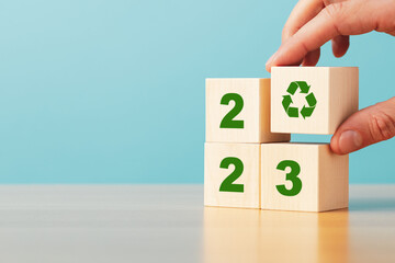 Hand putting wooden block with recycling icon on stack of blocks with numbers 2023 on blue...