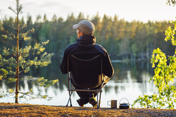 A man is sitting in a camping chair on a summer evening on the background of a forest lake. Object...